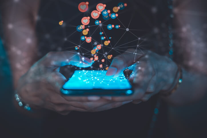 A person holds a smartphone glowing in the dark, with animated social media icons like hearts, likes, and emojis floating out, symbolizing online engagement, digital communication, and the impact of advertising.