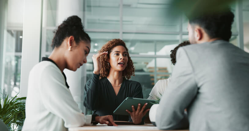 A woman leads a business discussion in a modern office, holding a tablet and speaking to colleagues. Two team members listen attentively, reflecting collaboration, strategy planning, and professional teamwork in a bright corporate setting.