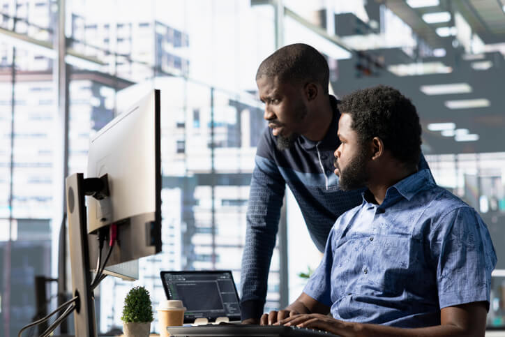 Two professionals work together at a computer, reviewing code and discussing cybersecurity strategies in an office.